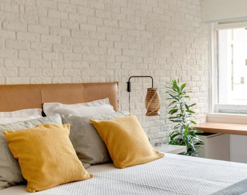 Quarto com parede de tijolinho branco pintado, cabeceira de couro caramelo e almofadas amarelas.